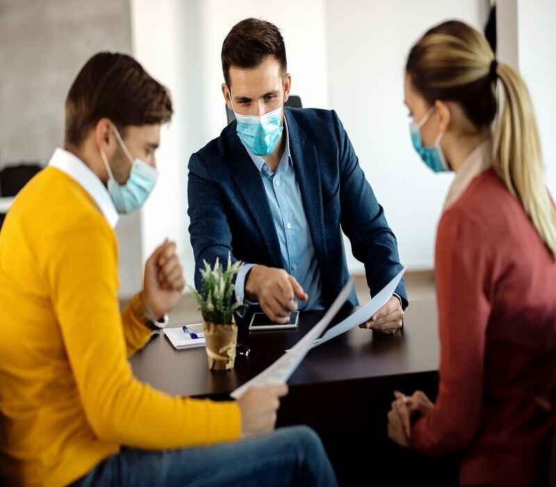 An insurance agent discussing policy details with two clients, all wearing masks for safety during a meeting.